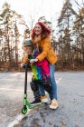 Encantada madre e hija montando patinete scooter en el estacionamiento mientras se divierten en fin de semana en otoño - foto de stock