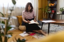 Artista donna adulta a corpo pieno seduta su cuscini vicino al tavolo con laptop e schizzo di disegno in sketchbook a casa — Foto stock