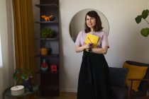 Felice donna con sketchbook e matita sorridente e guardando la fotocamera mentre in piedi vicino alla poltrona in studio creativo a casa — Foto stock