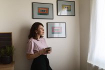 Мечтательная взрослая женщина с чашкой горячего напитка, глядя в окно и расслабляясь, опираясь на стену возле обрамленных изображений кассет дома — стоковое фото