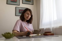 Сосредоточенный женский графический дизайнерский рисунок в альбоме эскизов, работая дистанционно над проектом за столом с чашкой кофе дома — стоковое фото