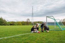 Padres ayudando a los hijos adolescentes a ponerse botas de fútbol mientras se preparan para jugar al fútbol en el campo verde - foto de stock