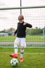 Delizioso adolescente in abbigliamento sportivo in piedi con palla sul campo di calcio vicino alla rete — Foto stock