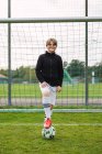 Восхитительный подросток в спортивной одежде стоит с мячом на футбольном поле рядом с сеткой и смотрит в камеру — стоковое фото