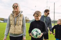 Веселая пара и сыновья-подростки в спортивной одежде собираются на зеленом футбольном поле, чтобы вместе играть в футбол на выходных — стоковое фото