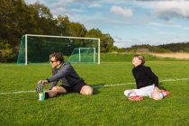 Alegre pai e adolescente em sportswear sentado esticando as pernas enquanto se prepara para jogar futebol no campo de futebol no verão — Fotografia de Stock