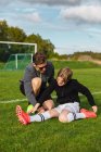 Adolescente em sportswear esticando as pernas antes do treinamento de futebol com a ajuda do treinador masculino no campo verde — Fotografia de Stock