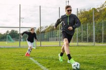 Веселий батько і підліток син в активному одязі грають у футбол, штовхаючи м'яч і бігаючи по полю — стокове фото