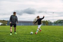 Веселый отец и сын-подросток в спортивной одежде играют в футбол, пиная мяч и бегая по полю — стоковое фото
