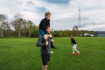 Jovem alegre chutando bola e jogando futebol em campo com pai e irmão durante o fim de semana — Fotografia de Stock