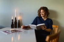 Adulto leitura feminina livro interessante enquanto sentado à mesa decorado com velas acesas em casa — Fotografia de Stock