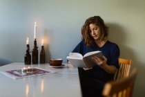Adult female reading interesting book while sitting at table decorated with burning candles at home — Stock Photo