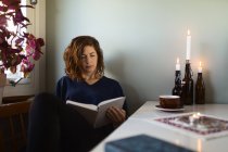 Erwachsene Frau liest interessantes Buch, während sie zu Hause am Tisch sitzt, der mit brennenden Kerzen dekoriert ist — Stockfoto