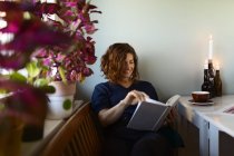 Donna adulta lettura interessante libro mentre seduto a tavola decorato con candele accese a casa — Foto stock