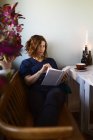 Adulte femelle lecture intéressant livre tandis que assis à la table décoré avec des bougies brûlantes à la maison — Photo de stock