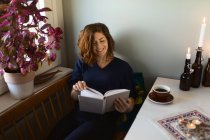 Erwachsene Frau liest interessantes Buch, während sie zu Hause am Tisch sitzt, der mit brennenden Kerzen dekoriert ist — Stockfoto