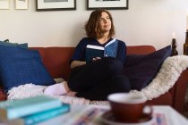 Щаслива жінка дивиться і мріє, сидячи на дивані і читаючи книгу вдома — стокове фото