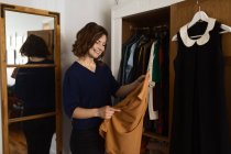 Glückliche erwachsene Frau lächelt und untersucht Kleidung, während sie in der Nähe des Kleiderschranks steht und das Outfit zu Hause auswählt — Stockfoto