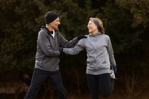 Positives altes Paar in Sportkleidung, das Arme und Beine ausstreckt, während es im Park trainiert und sich gegenseitig ansieht — Stockfoto