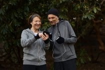 Sorrindo casal sênior em sportswear e fones de ouvido em pé juntos sob galhos de árvore e compartilhar telefone celular durante o treino de fitness — Fotografia de Stock