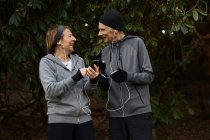 Улыбающаяся пожилая пара в спортивной одежде и наушниках, стоящая вместе под ветками деревьев и обменивающаяся мобильным телефоном во время тренировки — стоковое фото