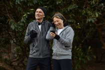 Sorrindo casal sênior em sportswear e fones de ouvido em pé juntos sob galhos de árvore e compartilhar telefone celular durante o treino de fitness — Fotografia de Stock