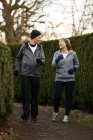 Corps entier d'un couple d'âge souriant portant des vêtements de sport et des gants et faisant du jogging entre des buissons verts dans le parc pendant l'entraînement physique — Photo de stock