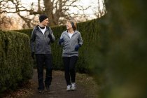Corps entier d'un couple d'âge souriant portant des vêtements de sport et des gants et faisant du jogging entre des buissons verts dans le parc pendant l'entraînement physique — Photo de stock