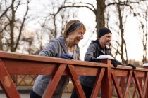 Smiling senior woman doing push up from wooden handrail with partner while having outdoor workout in autumn park — Stock Photo