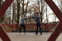 View from wooden fence of old positive couple warming up before workout in park with leafless trees — Stock Photo