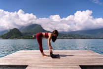 Side view of slender female preparing mat for doing yoga on wooden quay near lake in summer — Stock Photo