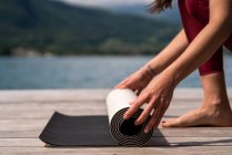 Side view of cropped unrecognizable slender female preparing mat for doing yoga on wooden quay near lake in summer — Stock Photo