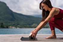 Side view of slender female preparing mat for doing yoga on wooden quay near lake in summer — Stock Photo