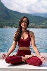 Donna serena seduta a Padmasana sul molo di legno e meditare con gli occhi chiusi mentre pratica yoga vicino al lago in estate — Foto stock