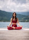 Серійна самиця сидить в Падмасані на дерев'яному пірсі і медитує закритими очима, практикуючи йогу біля озера влітку. — стокове фото
