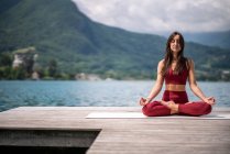 Серійна самиця сидить в Падмасані на дерев'яному пірсі і медитує закритими очима, практикуючи йогу біля озера влітку. — стокове фото
