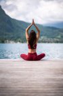 Rückansicht einer unkenntlich gelassenen Frau, die mit erhobenen Armen auf einem Holzsteg sitzt und meditiert, während sie im Sommer Yoga mit Mudra-Gesten in der Nähe des Sees praktiziert — Stockfoto