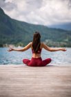 Back view of unrecognizable serene female sitting in Padmasana with arms up on wooden pier and meditating while practicing yoga with mudra gestures near lake in summer — Stock Photo