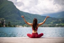 Vue arrière d'une femme sereine méconnaissable assise à Padmasana avec les bras levés sur une jetée en bois et méditant tout en pratiquant le yoga avec des gestes de mudra près du lac en été — Photo de stock