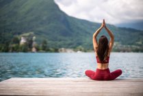 Vista posteriore di irriconoscibile donna serena seduta a Padmasana con le braccia alzate sul molo di legno e meditando durante la pratica dello yoga con gesti di mudra vicino al lago in estate — Foto stock