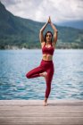 Femme flexible en vêtements de sport pratiquant le yoga tout en se tenant debout à Vrksasana avec les bras levés sur le quai près du lac regardant la caméra — Photo de stock