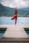 Flexible female in sportswear practicing yoga while standing in Vrksasana with raised arms on quay near lake looking at camera — Stock Photo
