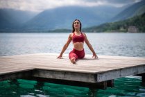Tranquillo femminile seduto a Hanumanasana sul molo di legno durante la pratica di yoga e stretching gambe vicino al lago in estate — Foto stock
