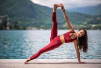 Flexible serene female in Utthita Vasishthasana practicing yoga and stretching legs on wooden pier near pond in summer — Stock Photo