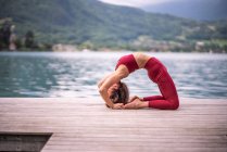 Vue latérale d'une femme flexible faisant du yoga à Kapotasana sur un quai en bois près du lac — Photo de stock
