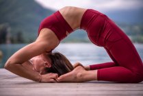 Vista laterale della femmina flessibile che fa yoga a Kapotasana sul molo di legno vicino al lago — Foto stock