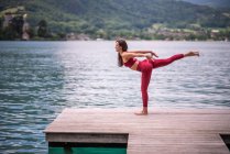 Бічний вид мирної жінки, яка балансує на нозі в Туладандасані під час практики йоги на дерев'яному пірсі біля ставка, дивлячись у бік — стокове фото