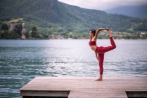 Vista laterale di pacifico equilibrio femminile sulla gamba a Natarajasana durante la pratica dello yoga sul molo di legno vicino allo stagno guardando altrove — Foto stock