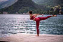 Seitenansicht einer friedlichen Frau, die in Tuladandasana auf einem Bein balanciert, während sie Yoga auf einem hölzernen Pier in der Nähe eines Teiches praktiziert, der wegschaut — Stockfoto