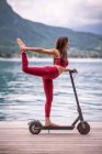 Vista lateral de la mujer flexible encantada en ropa deportiva equilibrio en Natarajasana en scooter eléctrico mientras practica yoga en el muelle de madera y mirando hacia otro lado - foto de stock